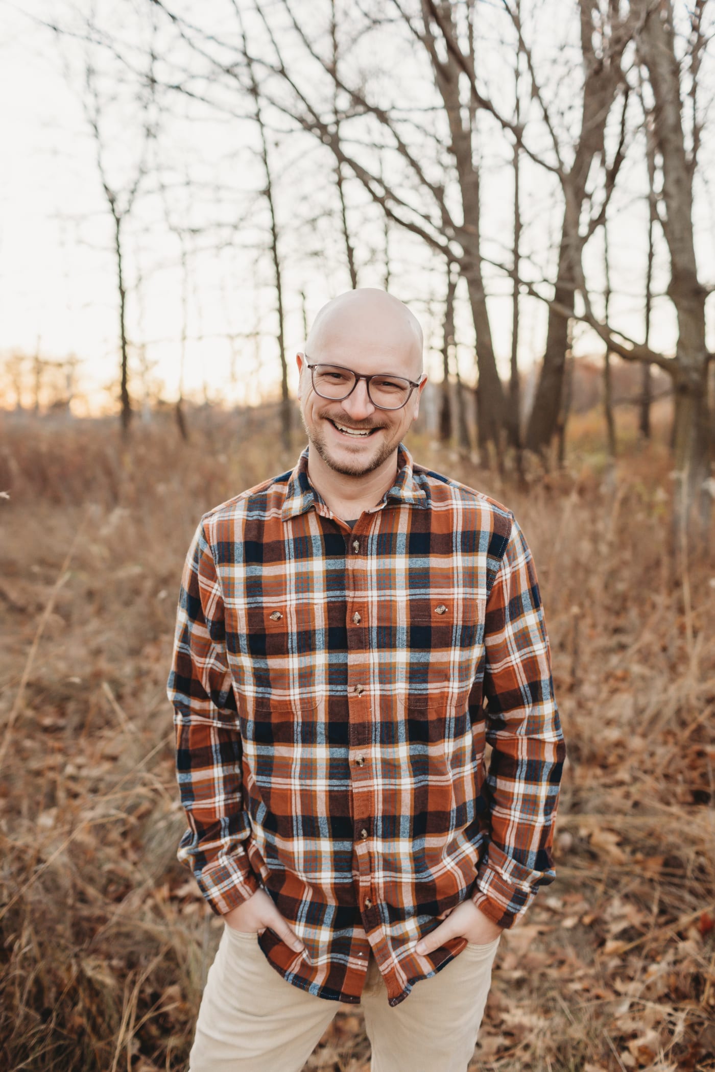 A portrait of Brian Muenzenmeyer. Brian is a 30-somthing bald white man wearing a plaid shirt, grey glasses, and a five o'lock shadow. Photo taken by Samantha Brinda Photography